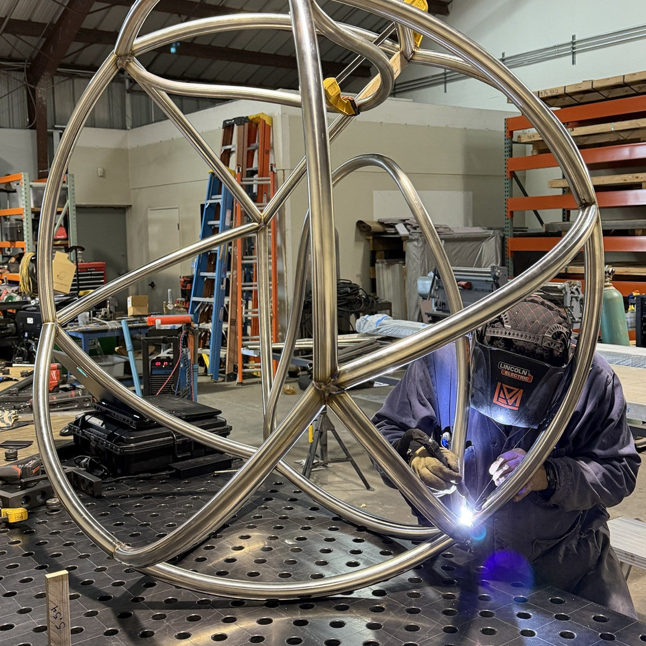 A welder in a helmet TIG welds a joint on the stainless steel Kora frame, the arc glowing blue-white