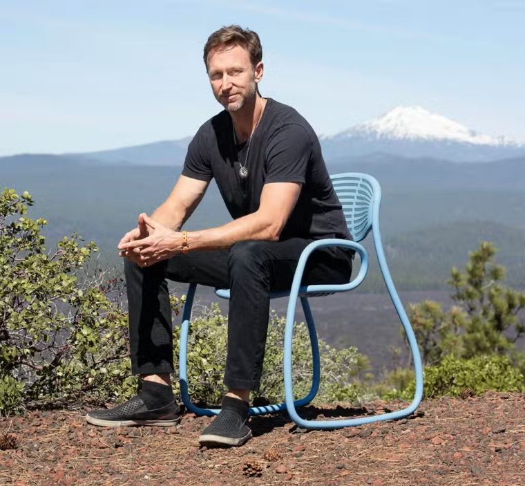 Jules Henry seated in a blue Vibe chair on a ridge, a snow-capped peak behind him