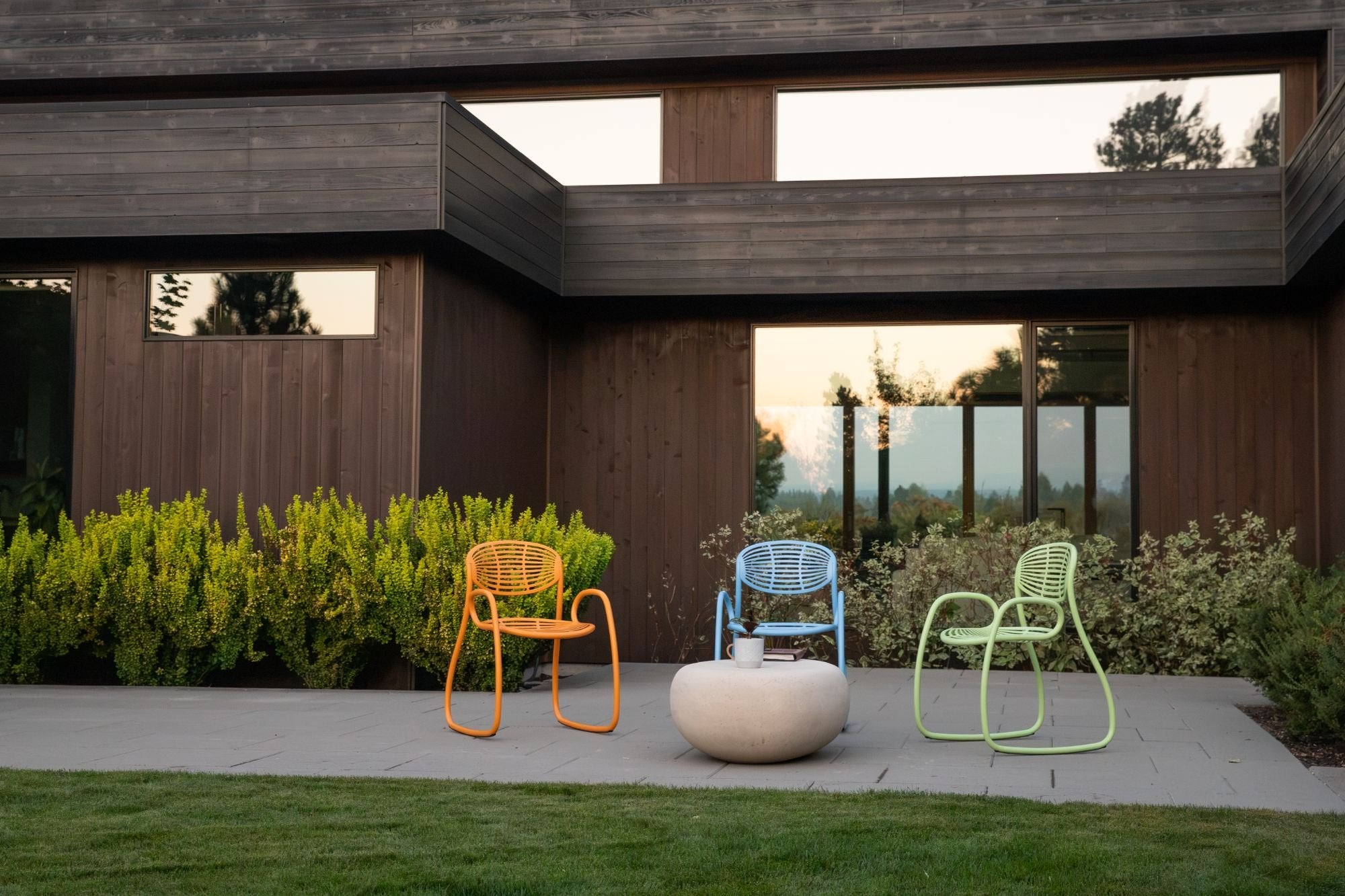 Three Vibe chairs, orange, blue and lime green, on a stone patio in front of a dark wooden house at sunset