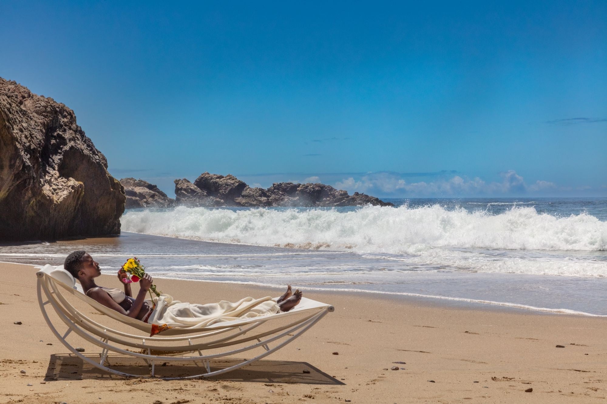 A woman resting in a white Volo lounge on a beach as a wave breaks behind her