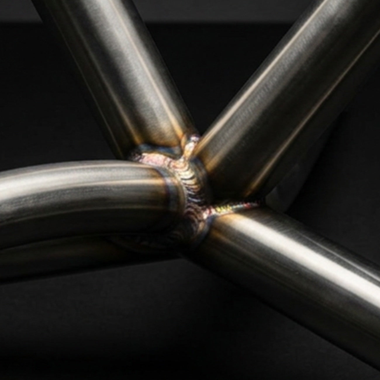 A close-up of a TIG-welded stainless steel joint where several tubes meet, lit in a dark studio